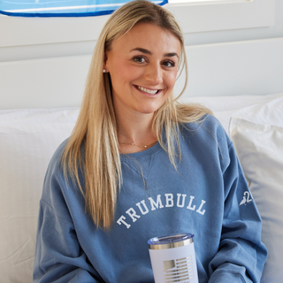 Woman wearing a blue 'TRUMBULL' sweatshirt holding a white tumbler with a blue lid.