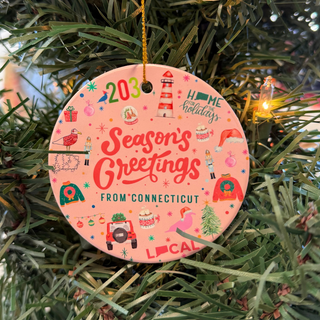 A round ceramic holiday ornament with a 'Season's Greetings from Connecticut' text, featuring holiday-themed illustrations in red, green, and yellow colors, hanging on a Christmas tree.